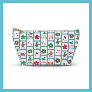 May include: A rectangular cosmetic bag with a gold zipper. The bag is patterned with white tiles featuring red, green, and black symbols and characters. The bag's design includes the words "Crak", "Bam", and "Joker".