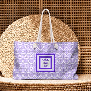 May include: A tote bag with a purple and white geometric pattern. The bag has white rope handles and a white square with the text "CRACK BAM DOT" in the center. The bag is sitting on a woven surface.