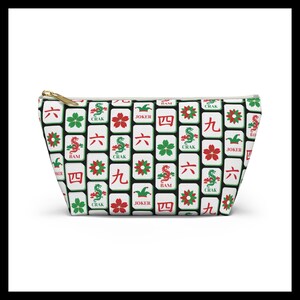 May include: A white cosmetic bag with a gold zipper, featuring a repeating pattern of Mahjong tiles in red, green, and black. The tiles display various symbols and characters, including dragons, flowers, and numbers.