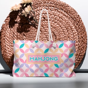 May include: A large tote bag with a geometric pattern in pastel colors of pink, blue, yellow, and purple. The bag has white rope handles and the word "MAHJONG" in turquoise letters. The background is a woven basket.