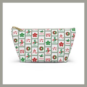 May include: A white zippered pouch with a repeating pattern of Mahjong tiles in red, green, and black. The tiles feature various symbols and characters, including dragons, flowers, and numbers. The zipper is gold.