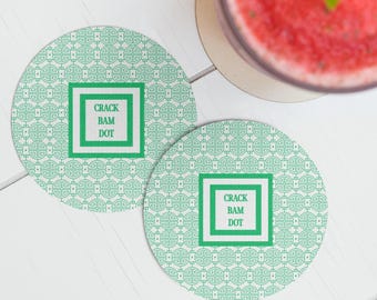 May include: Two round coasters with a green and white pattern and the text "CRACK BAM DOT" in a green square. The coasters are on a white wooden surface, with a glass of red liquid in the background.