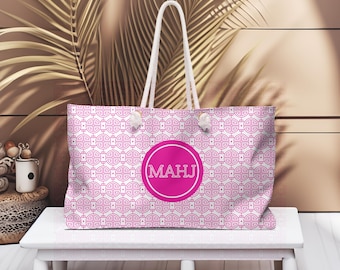 May include: A pink and white tote bag featuring a geometric pattern. It has white rope handles and a large pink circle with the letters "MAHJ". The bag is on a white table. The bag's design is visible.