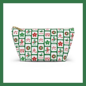 May include: A white zippered pouch featuring a repeating pattern of Mahjong tiles in green, red, and white. The tiles display various symbols and characters, including flowers, dragons, and numbers. The pouch has a gold-colored zipper.