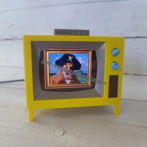 Op de afbeelding: Een felgele televisie-vormige lamp met een scherm waarop een piraat in een goudkleurige lijst te zien is. Het apparaat heeft twee lichtblauwe ronde knoppen aan de rechterkant, een donkerbruine verticale ventilatieopening en een grijs bovendeel.
