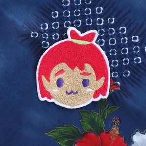 May include: A colorful embroidered patch featuring a cartoon character with red hair, a green bow, and a tan face. The patch is on a dark blue fabric with a floral design and a woven straw mat.