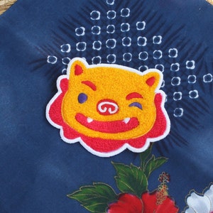 May include: A colorful, embroidered lion patch with a winking expression. The patch is yellow, red, and white, and is set against a dark blue patterned fabric with floral accents. The patch is likely for clothing or accessories.