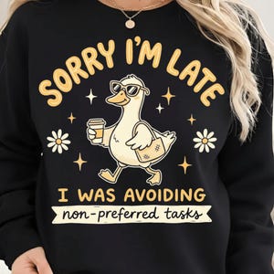 Puede incluir: Sudadera negra con un gráfico humorístico. El diseño presenta un pato de dibujos animados con gafas de sol, llevando una taza de café y una bolsa, con el texto "SORRY I'M LATE" y "I WAS AVOIDING non-preferred tasks."