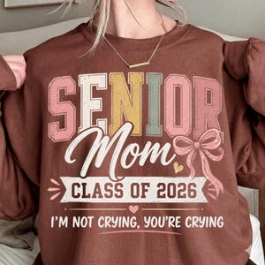 Può includere: Una felpa marrone con la scritta "SENIOR Mom CLASS OF 2026" in un design retrò colorato. Il testo "I'M NOT CRYING, YOU'RE CRYING" è sotto. Il design include un fiocco rosa e un cuore.