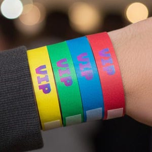 May include: A wrist with four colorful VIP wristbands. The bands are yellow, green, blue, and red. The word "VIP" is printed on each band in purple. The person is wearing a dark gray sweatshirt.