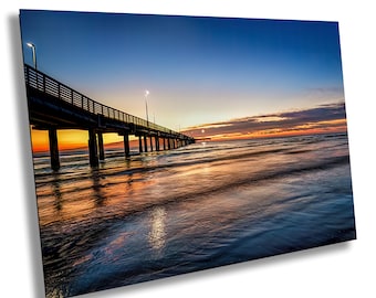 May include: A wooden pier extends out into the ocean at sunset. The water is calm and reflects the colors of the sky, which is a mix of blue, orange, and pink. The pier is silhouetted against the sky.
