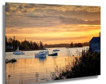 May include: A scenic landscape print featuring a harbor at sunset. Several boats are docked in calm water, reflecting the warm hues of the sky. The sky is filled with golden and orange tones, with a few clouds. A small building is on the right.