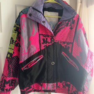 May include: A black, pink, and gray patterned jacket with a zipper closure and a snap button. The jacket has two zippered pockets on the front.