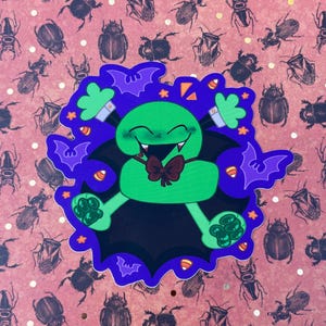 May include: A green cartoon vampire sticker with a black cape, purple bat wings, and a red bow tie. The sticker has a smiling face with fangs and is surrounded by candy corn and stars. The background is a pink pattern with insect illustrations.