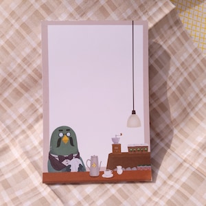 Puede incluir: Un personaje de pájaro verde con bigote y gafas está sentado en una mesa con una cafetera, un molinillo de café y una taza de café. El pájaro lleva una corbata de moño y sostiene una servilleta. Hay una luz colgando del techo sobre la mesa.