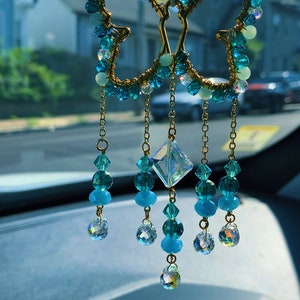 May include: A gold butterfly-shaped car charm with blue and green crystal beads hanging from it. The charm is hanging from the rearview mirror of a car.