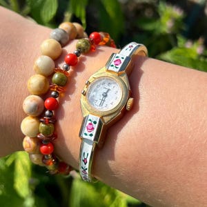 Peut inclure: Une montre analogique dorée avec un cadran blanc et des motifs floraux en émail sur le bracelet, accompagnée d'un bracelet composé de perles rondes beiges, vertes et rouges avec de petites entretoises métalliques.