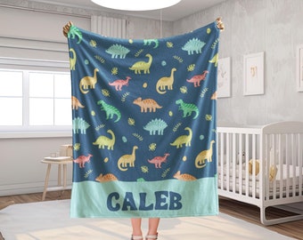 May include: A navy blue blanket featuring colorful dinosaur illustrations. The blanket has a light blue border with the name "CALEB" in white block letters. The dinosaurs are in shades of yellow, green, orange, and pink.