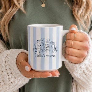 May include: A white ceramic mug featuring vertical light blue stripes and a dark blue line art illustration of a wildflower bouquet. The text "Stella's mama" is printed in a dark blue script font centred below the floral design.
