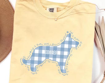 May include: A pale yellow t-shirt featuring a blue and white gingham patterned silhouette of a German Shepherd dog. The name COOPER is printed in white inside the silhouette, surrounded by the repeating text Life Is Better With A German Shepherd.