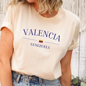 May include: Cream-colored t-shirt with the word "VALENCIA" in navy blue, a horizontal line, and the word "VENEZUELA" below it. A small Venezuelan flag is between the line and the word "VENEZUELA". The shirt has rolled-up sleeves.