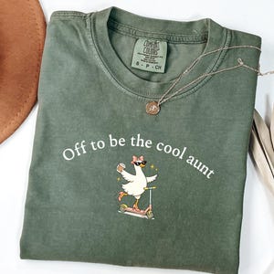 May include: An olive green short-sleeve t-shirt with a crew neckline. The shirt features a graphic of a white goose wearing sunglasses, a pink bow, and pink heels, riding a scooter while holding a coffee cup. The text reads, Off to be the cool aunt.
