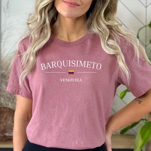 May include: A dusty rose t-shirt with the word "BARQUISIMETO" in white capital letters, with a horizontal line and the Venezuelan flag below it. The word "VENEZUELA" is printed below the flag. The shirt is short-sleeved.
