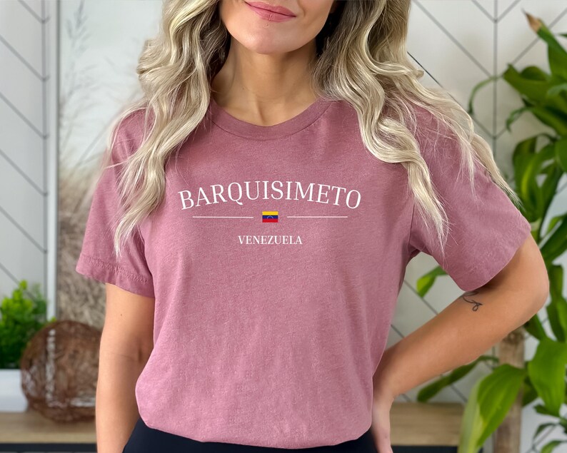May include: A dusty rose t-shirt with the word "BARQUISIMETO" in white capital letters, with a horizontal line and the Venezuelan flag below it. The word "VENEZUELA" is printed below the flag. The shirt is short-sleeved.