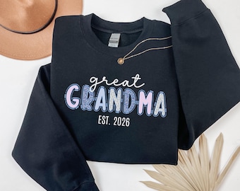 May include: Black sweatshirt with the words "great GRANDMA" in white, blue, and pink lettering. The text "EST. 2026" is below the main text. A gold necklace and a brown hat are visible in the background.