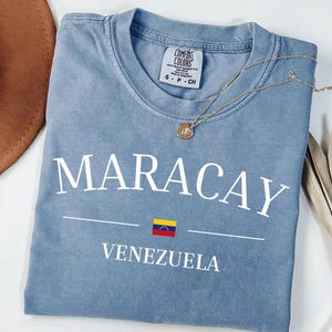 Op de afbeelding: Een blauw T-shirt met korte mouwen voorzien van de witte tekst MARACAY en VENEZUELA met een kleine Venezolaanse vlag ertussen. Het shirt heeft een ronde hals en een Comfort Colors merklabel zichtbaar bij de binnenkant van de kraag.