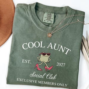 May include: An olive green t-shirt with the text "COOL AUNT" and "Social Club" printed in white. The shirt features a cartoon frog wearing sunglasses and pink high heels. A gold necklace and a brown hat are also visible.