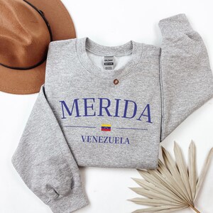 Pu&ograve; includere: Felpa grigia con il testo "MERIDA VENEZUELA" in lettere blu. Una piccola bandiera venezuelana &egrave; stampata tra le parole.