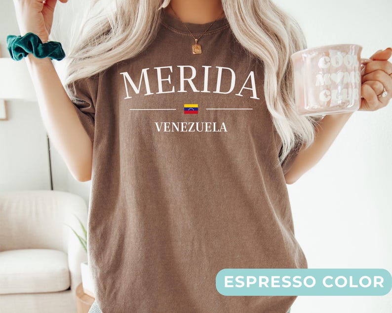 Op de afbeelding: Een espresso-kleurig t-shirt met de witte tekst MERIDA gecentreerd boven een kleine Venezolaanse vlag en het woord VENEZUELA. Het t-shirt heeft een ronde hals en een relaxte pasvorm met een eenvoudig grafisch ontwerp op de borst gedrukt.