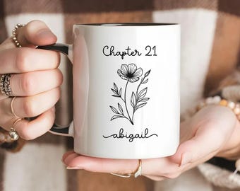 May include: A white ceramic mug with a black interior and handle. The front features the black text "Chapter 21" at the top, a black line-art illustration of a single flower in the center, and the name "abigail" in black script at the bottom.