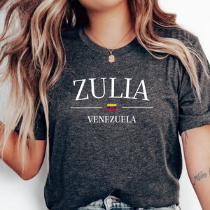 May include: Dark gray t-shirt with the word "ZULIA" in white, above "VENEZUELA" in white. A horizontal line separates the words, with the Venezuelan flag in the middle. The shirt is a crew neck.