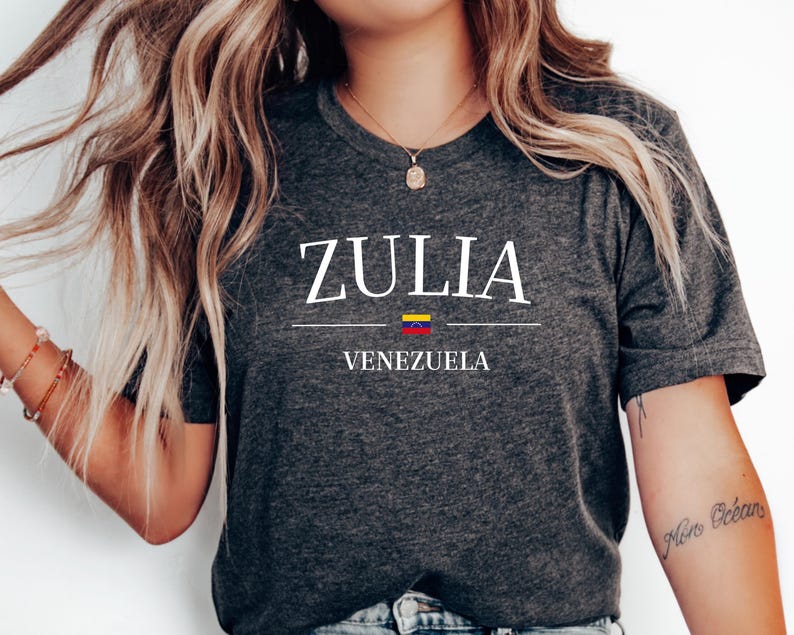 May include: Dark gray t-shirt with the word "ZULIA" in white, above "VENEZUELA" in white. A horizontal line separates the words, with the Venezuelan flag in the middle. The shirt is a crew neck.