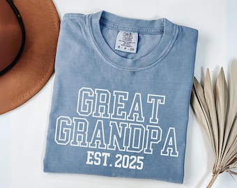 May include: A light blue Comfort Colors t-shirt with the words "GREAT GRANDPA" and "EST. 2025" in white block letters. The shirt is folded and lies on a white surface, with a brown hat and dried plant in the background.