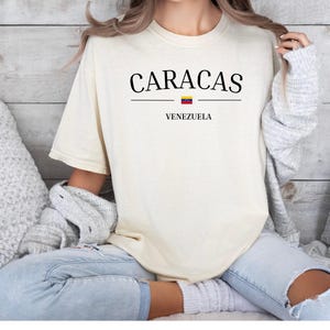 Op de afbeelding: Een cr&egrave;mekleurig t-shirt met korte mouwen voorzien van de zwarte tekst CARACAS gecentreerd boven een kleine horizontale lijn met het icoon van de Venezolaanse vlag en het woord VENEZUELA in het zwart onder de lijn op de voorzijde.
