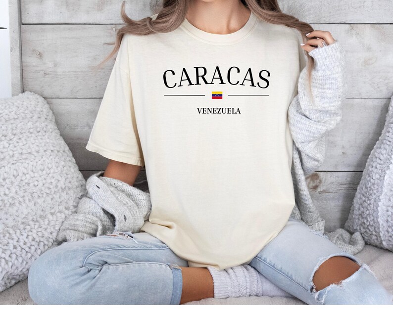 Op de afbeelding: Een cr&egrave;mekleurig t-shirt met korte mouwen voorzien van de zwarte tekst CARACAS gecentreerd boven een kleine horizontale lijn met het icoon van de Venezolaanse vlag en het woord VENEZUELA in het zwart onder de lijn op de voorzijde.