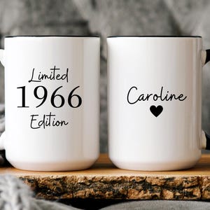 May include: Two white ceramic mugs with black handles and rims. One mug reads "Limited 1966 Edition" in black script. The other mug has the name "Caroline" and a black heart. Both mugs are on a wooden surface.