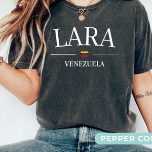 Op de afbeelding: Een antracietgrijs t-shirt met korte mouwen en de witte tekst LARA gecentreerd boven een kleine horizontale lijn, een miniatuurvlag van Venezuela en het woord VENEZUELA eronder. Het shirt heeft een vintage gewassen afwerking en een ronde hals.