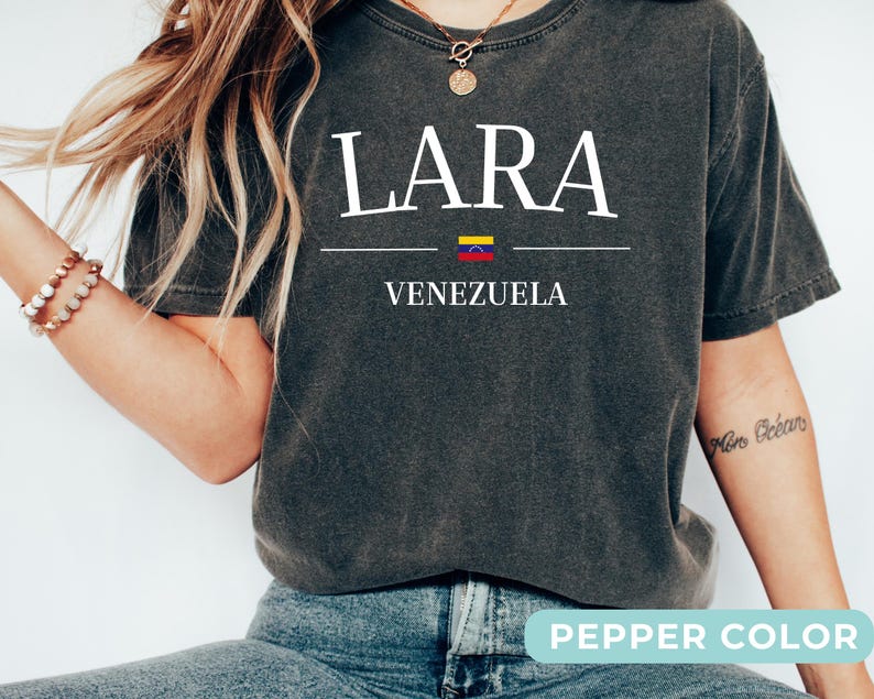 Op de afbeelding: Een antracietgrijs t-shirt met korte mouwen en de witte tekst LARA gecentreerd boven een kleine horizontale lijn, een miniatuurvlag van Venezuela en het woord VENEZUELA eronder. Het shirt heeft een vintage gewassen afwerking en een ronde hals.