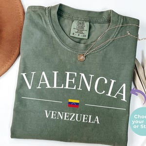 Op de afbeelding: Een olijfgroen T-shirt met korte mouwen voorzien van de witte tekst VALENCIA boven een kleine Venezolaanse vlag en het woord VENEZUELA. Het shirt heeft een ronde hals en een Comfort Colors merklabel aan de binnenkant met maat small.