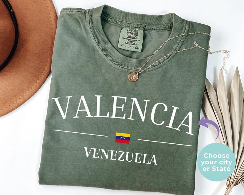 Op de afbeelding: Een olijfgroen T-shirt met korte mouwen voorzien van de witte tekst VALENCIA boven een kleine Venezolaanse vlag en het woord VENEZUELA. Het shirt heeft een ronde hals en een Comfort Colors merklabel aan de binnenkant met maat small.