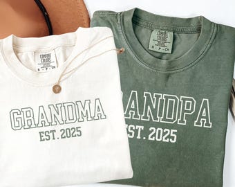 May include: Two crewneck t-shirts, one cream and one olive green, featuring the text GRANDMA EST. 2025 and GRANDPA EST. 2025 in a white collegiate font. Both shirts have a Comfort Colors label at the inner neckline and a size S tag.
