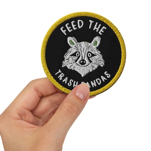 Puede incluir: Un parche bordado negro y amarillo con la cara de un mapache y el texto "Feed the Trash Pandas".