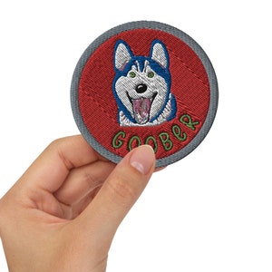 Puede incluir: Un parche redondo bordado de color rojo con una ilustración de un perro husky azul y blanco y la palabra "GOOBER" en letras verdes.