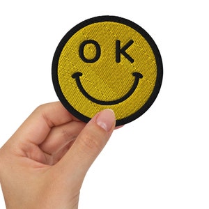 Könnte beinhalten: Ein gelber Smiley-Patch mit schwarzem Rand und dem Wort "OK" in schwarzen Buchstaben.