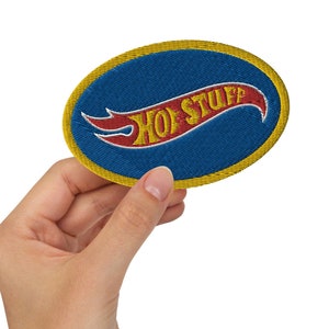 May include: A blue and yellow oval-shaped patch with the words "Hot Stuff" in red and yellow lettering. The patch is embroidered and has a yellow border.