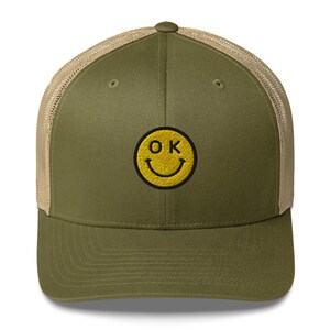May include: A green and tan trucker hat with a yellow smiley face embroidered on the front. The smiley face has the letters "OK" inside of it.